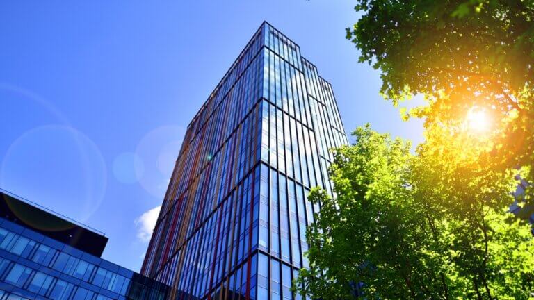 A glass building with a blue sky and rays of sun through trees.